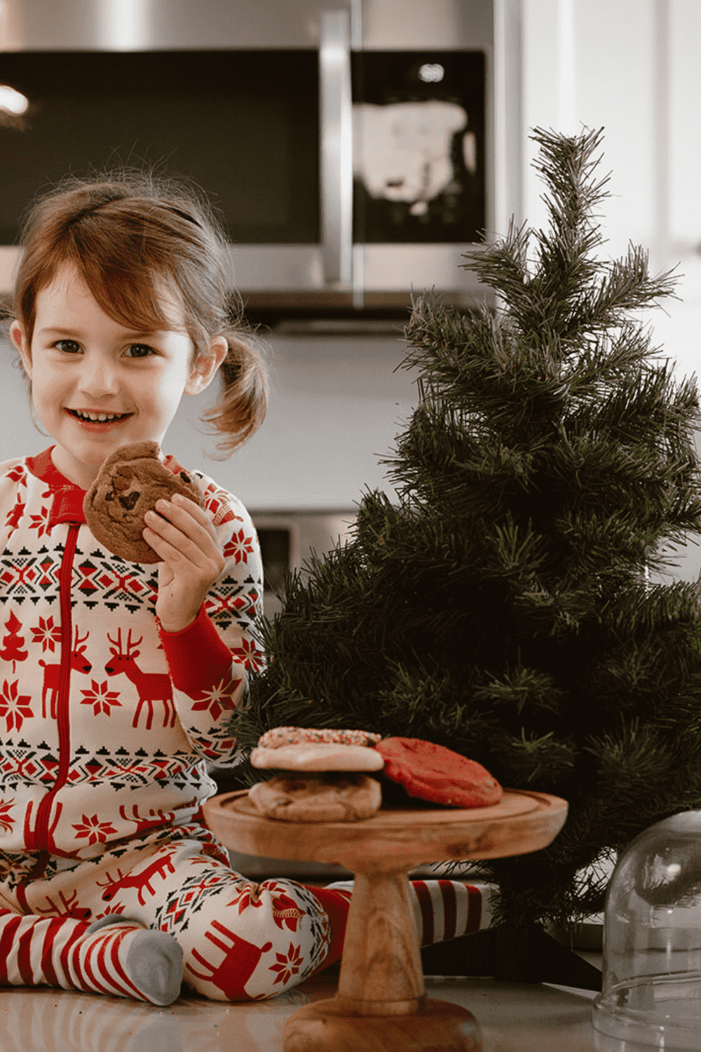 A little girl dressed in holiday pyjamas holding a chocolate chip cookie, sitting beside a Christmas tree and a tray of cookies.