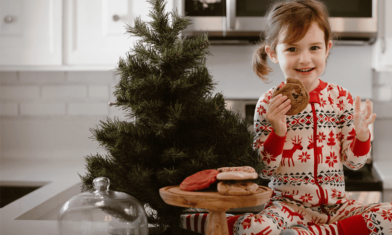 A little girl dressed in holiday pyjamas holding a chocolate chip cookie, sitting beside a Christmas tree and a tray of cookies.