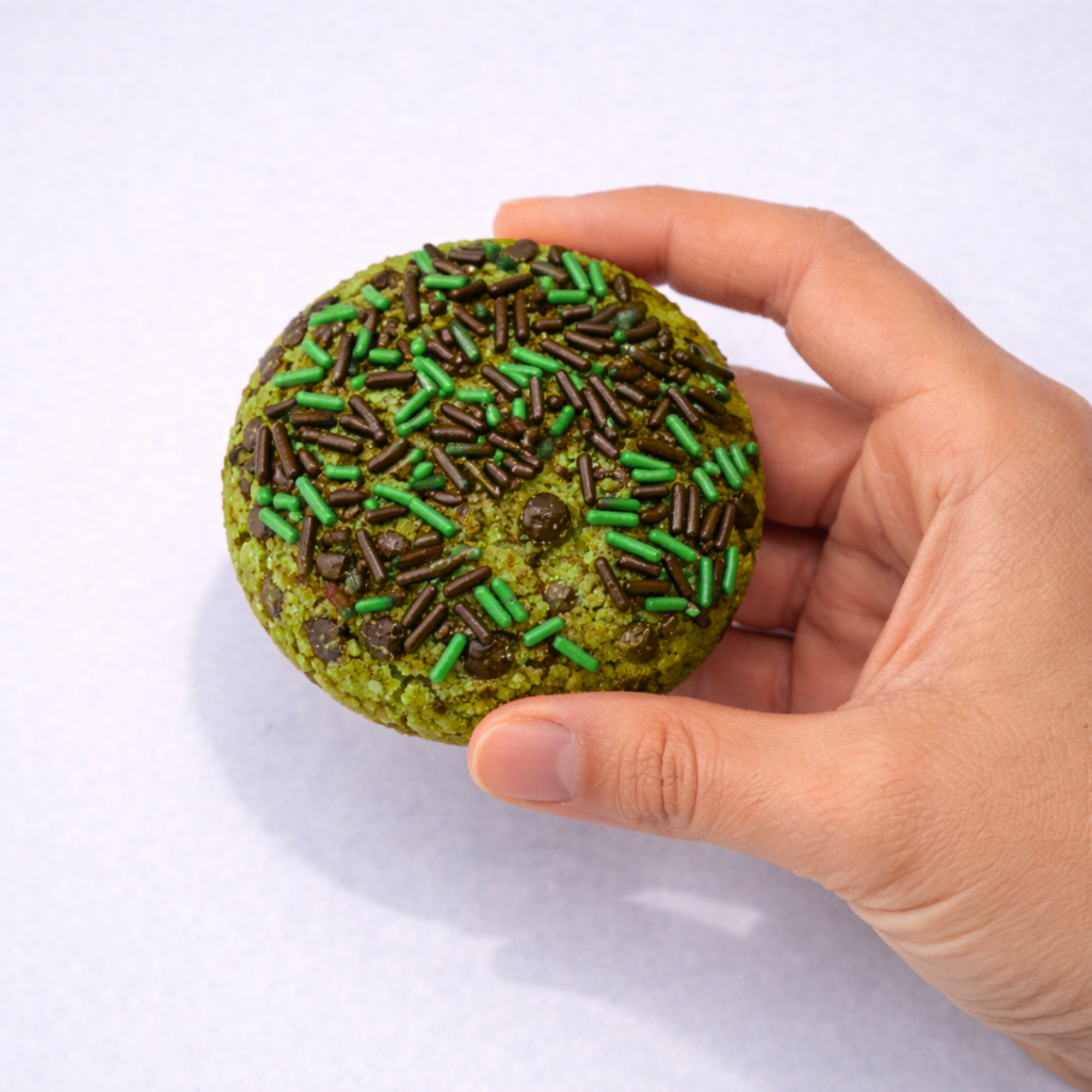 Hand holding a green cookie with chocolate chips and green sprinkles on a white background