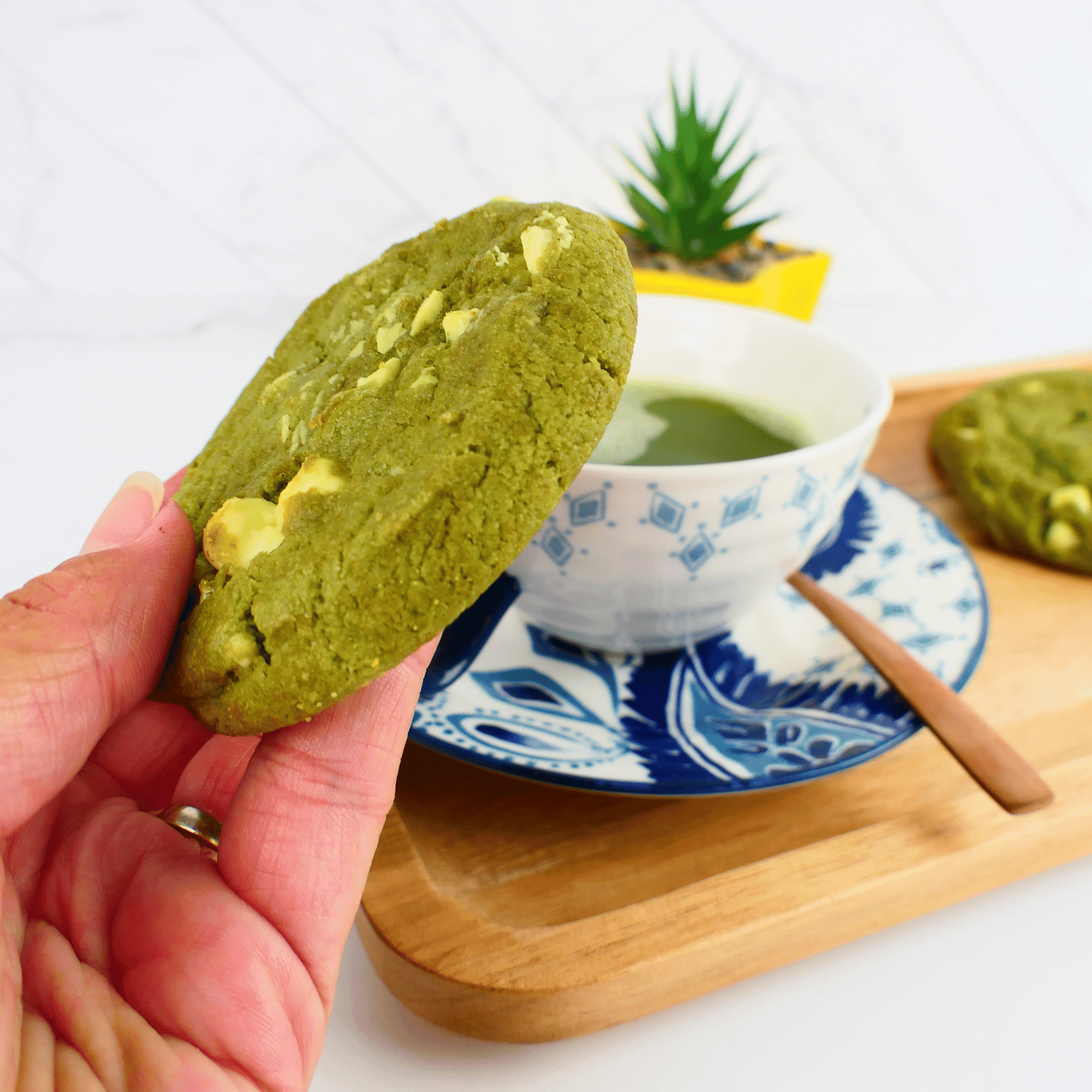 Green matcha cookie held by a hand with a cup of green tea on a wooden tray.