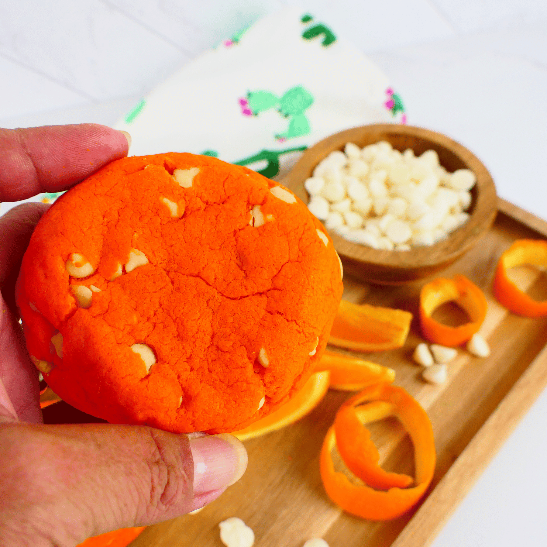 Orange cookie held by a hand, on a wooden tray with a bowl of yogurt chips and orange peels.