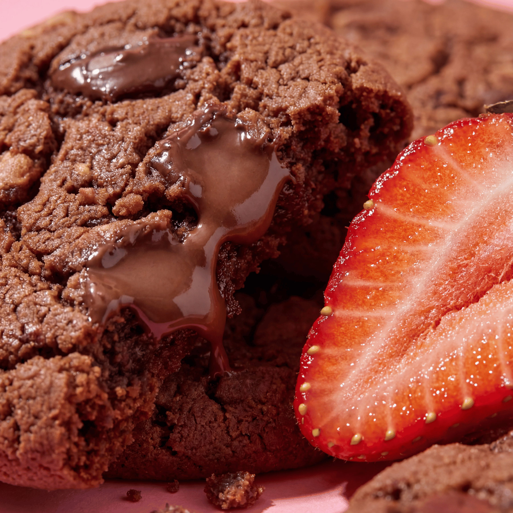 Chocolate cookie with a gooey center next to a sliced strawberry on a pink surface