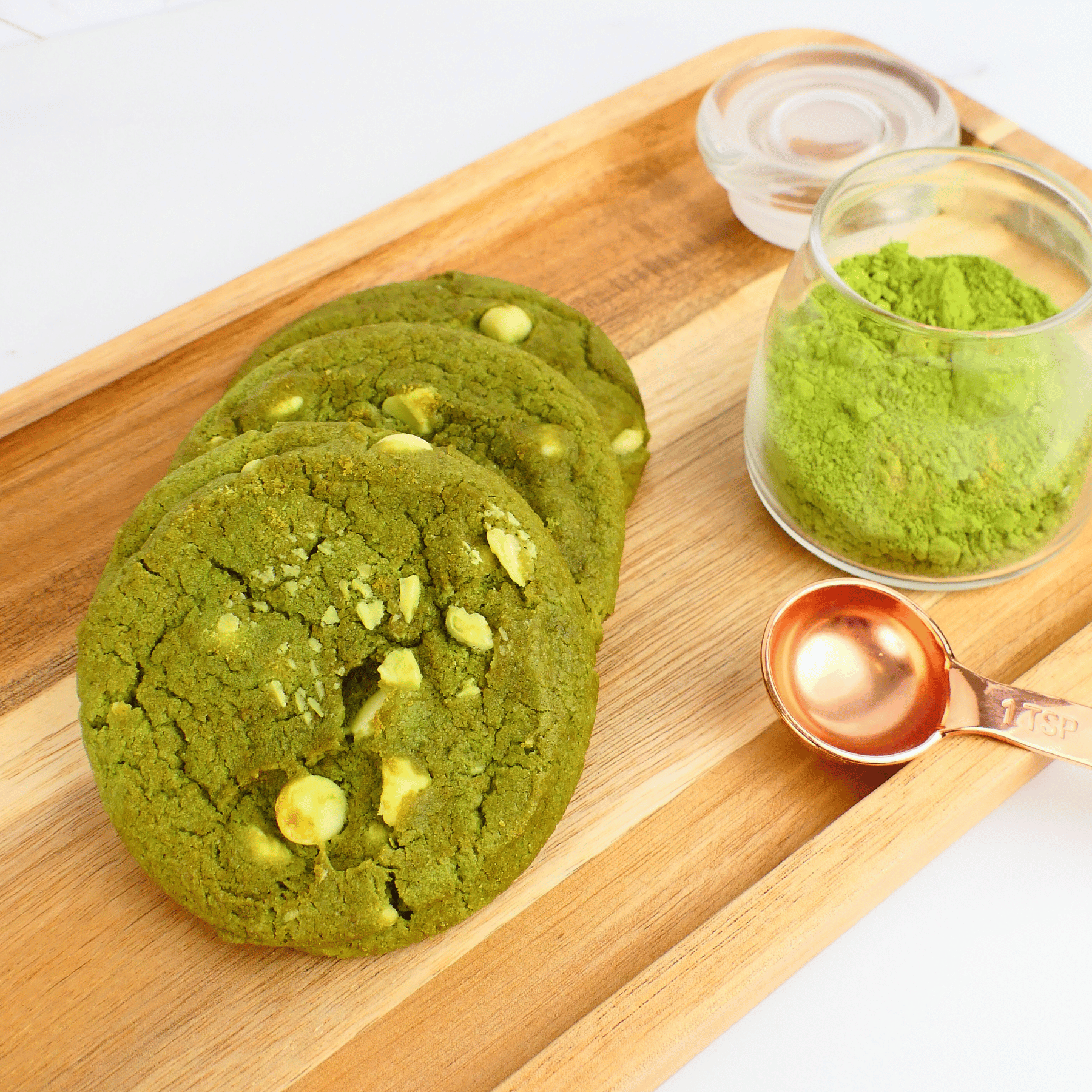 Green cookies with white chocolate chips on a wooden board with a small container of matcha powder and a measuring spoon.